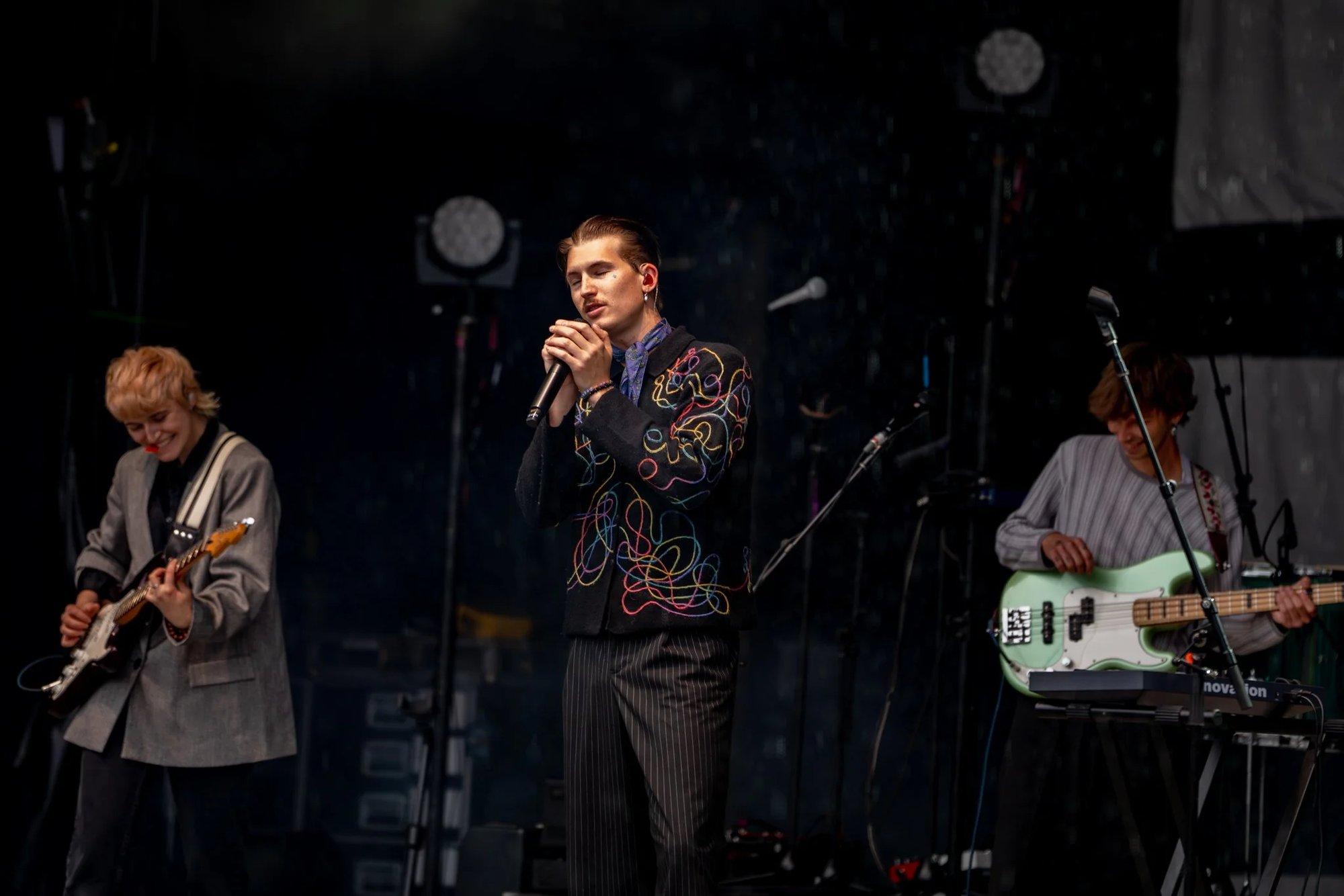 Singer cradling the mic in an embroidered jacket, bandmates at guitar and bass in the shadows