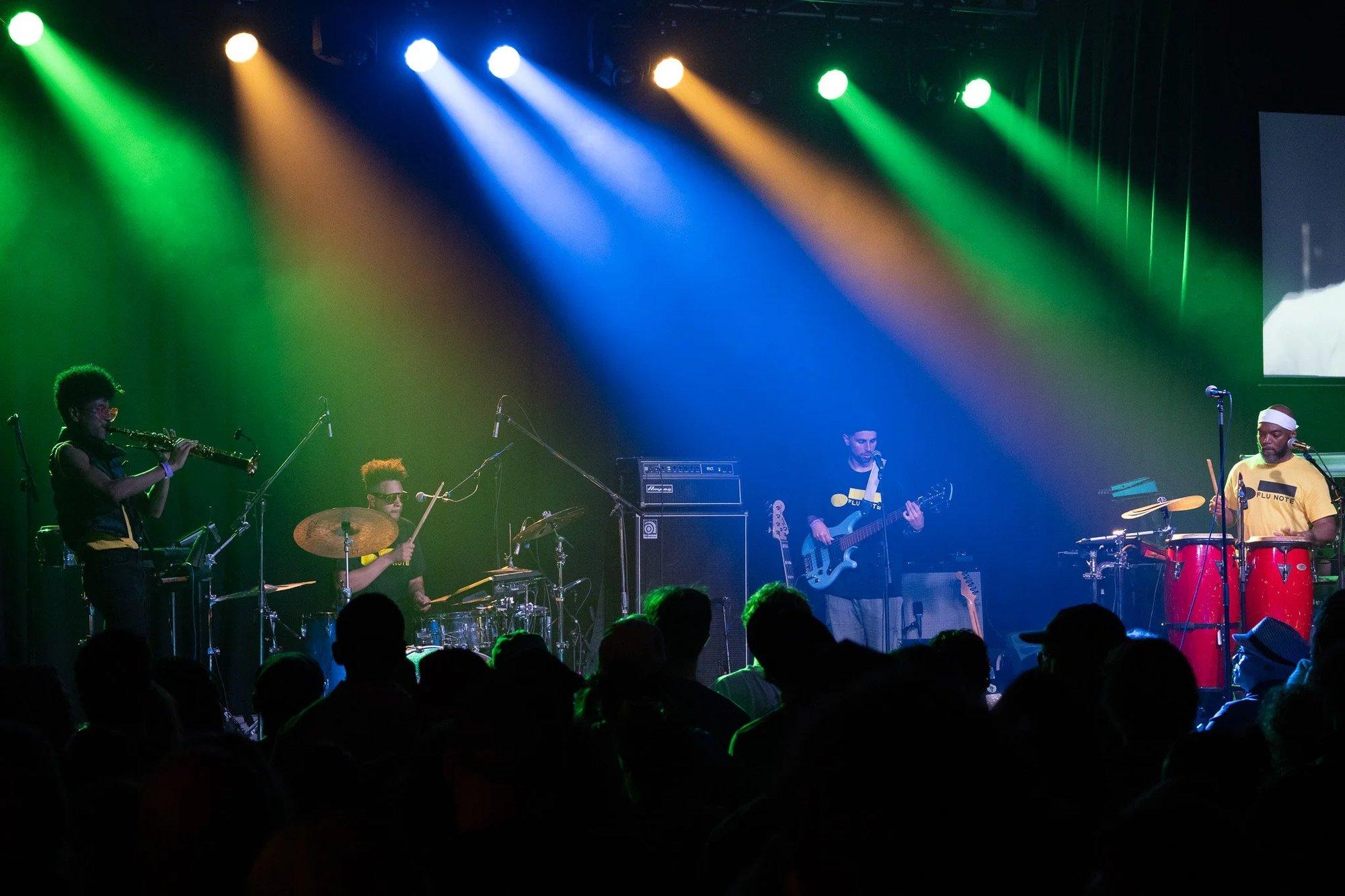 Five-piece band with sax and congas under fanned green, blue, and amber beams above the crowd