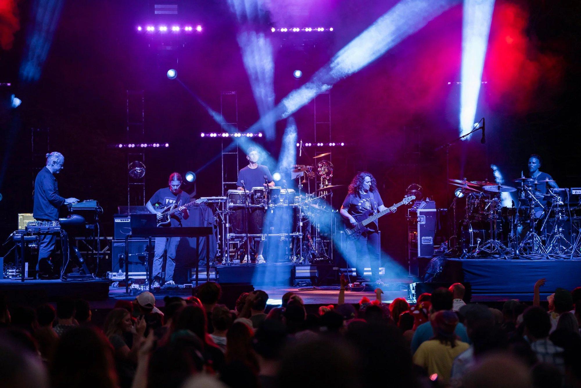 Full stage bathed in purple haze as white spotlight beams cross over a silhouetted crowd