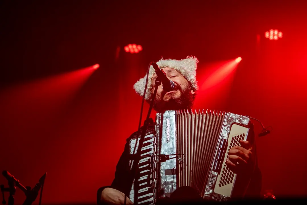 Accordion player in a white fur hat singing into the mic, engulfed in deep red light