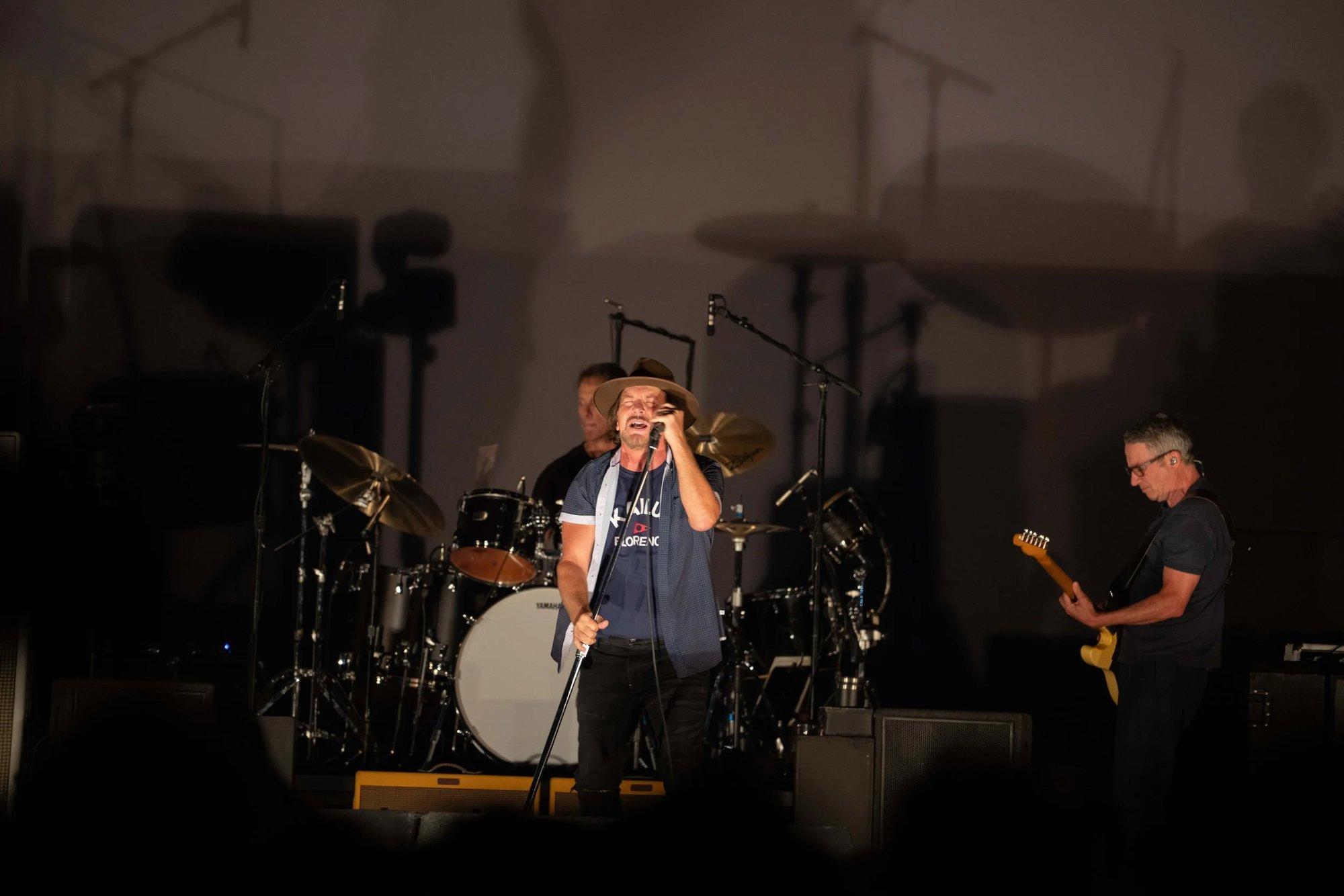 Eddie Vedder of Pearl Jam singing into the mic under a single warm spotlight, band in shadow behind him